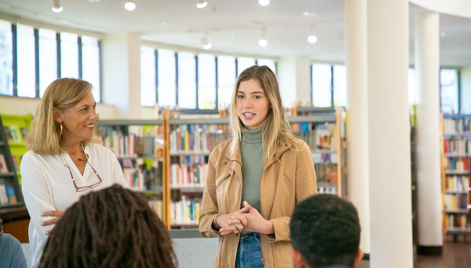 Student presenting to class in library while teacher watches