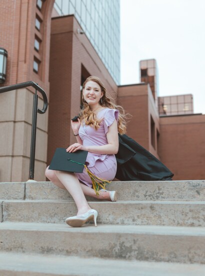 Graduate sitting on stairs in front of college with cap and gown